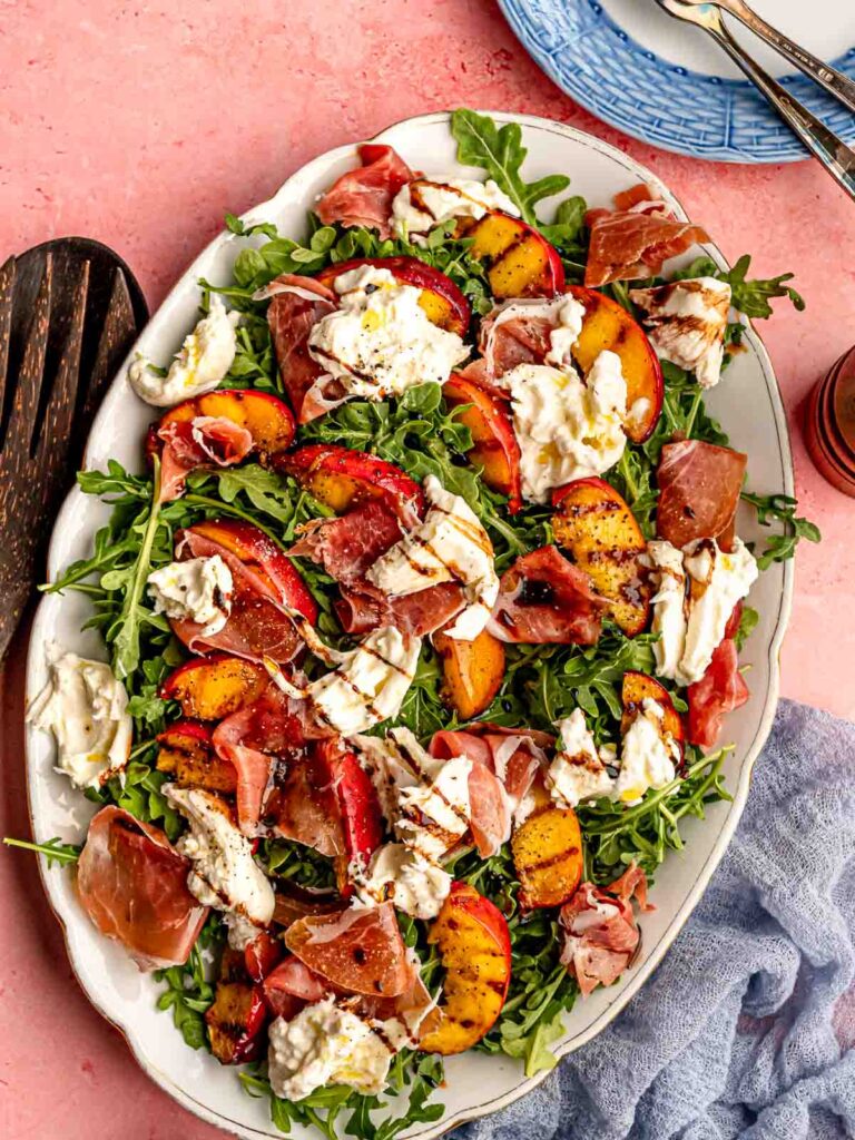 One of our favorite summer recipes, a vibrant plate of grilled peach salad featuring juicy, caramelized peach slices, fresh mixed greens, crumbled feta cheese, and toasted pecans, all drizzled with a light balsamic vinaigrette.