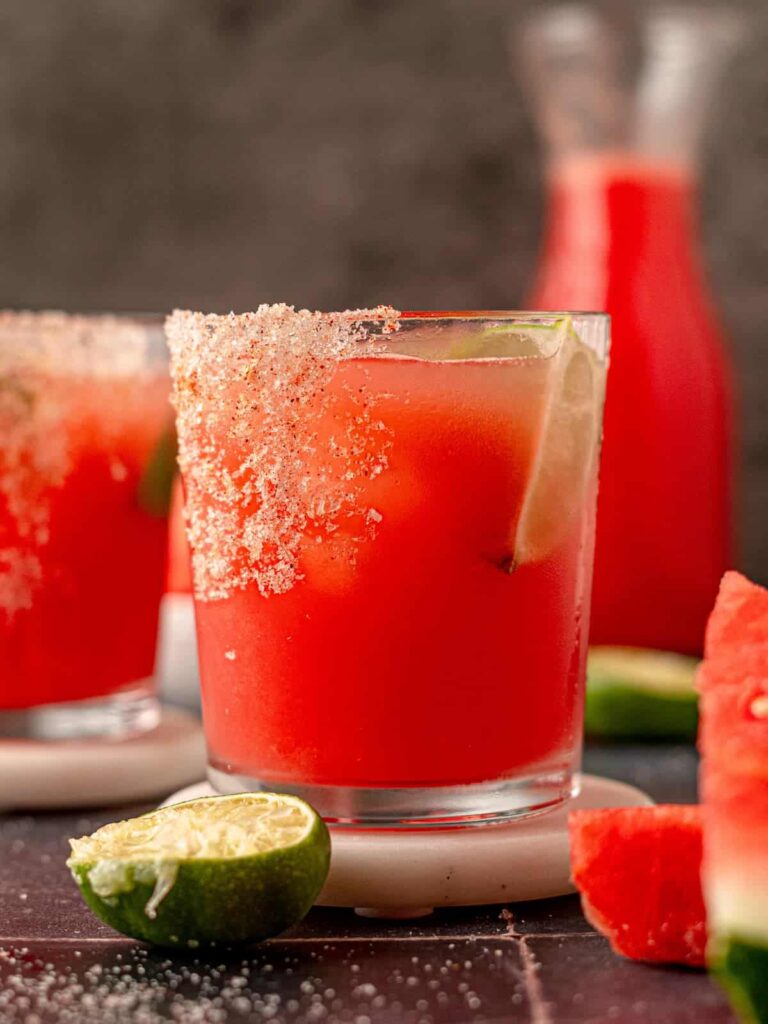 One of our favorite summer recipes, refreshing spicy watermelon margaritas served in salt-rimmed glasses, garnished with lime wedges and jalapeño slices, alongside a bowl of fresh watermelon chunks.